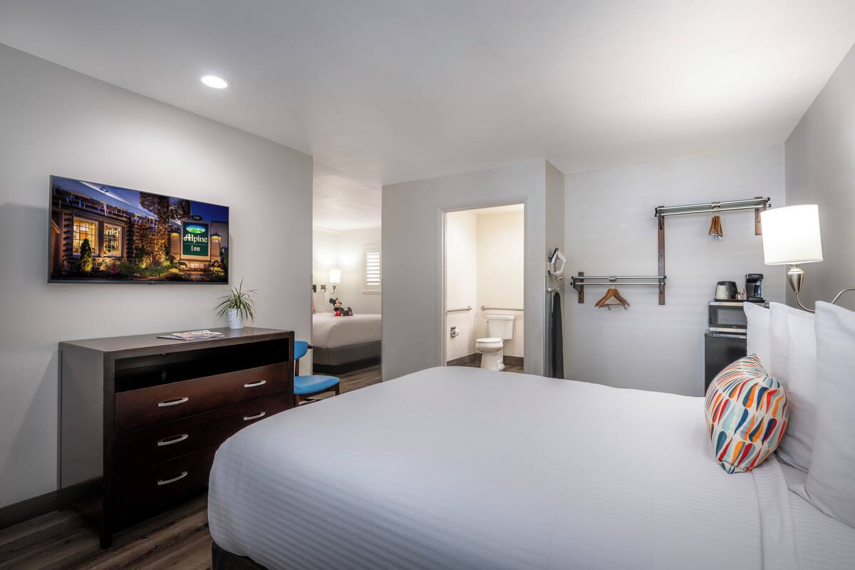 Ein Hotelzimmer mit Bett, Schreibtisch mit Schubladen, Stuhl, an der Wand montiertem Fernseher sowie einem sichtbaren Badezimmer durch eine geöffnete Tür und einem Hängehandtuchhalter.