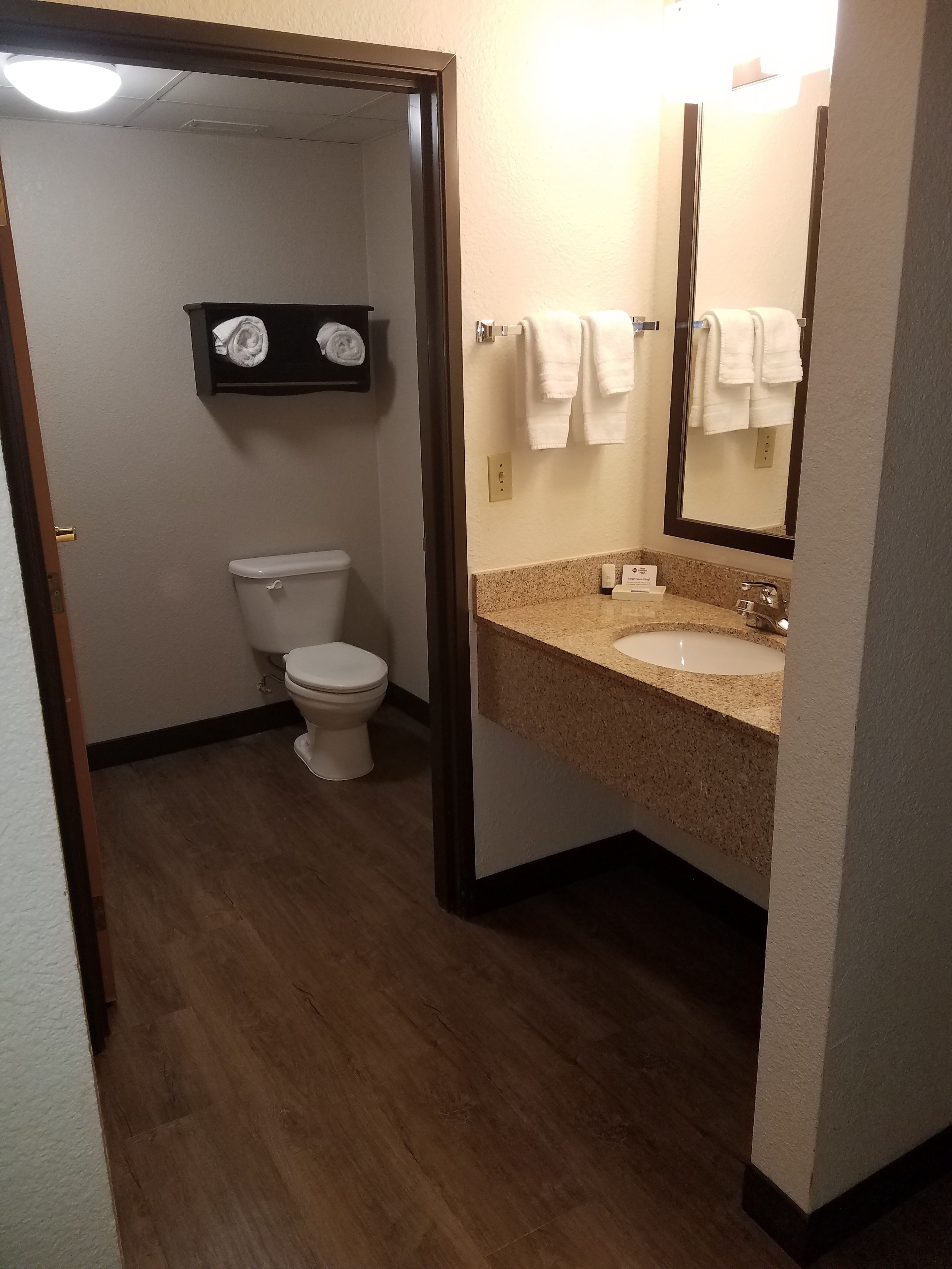 A bathroom with a toilet, sink, and mirror. There is a towel rack above the sink and a shelf with towels.