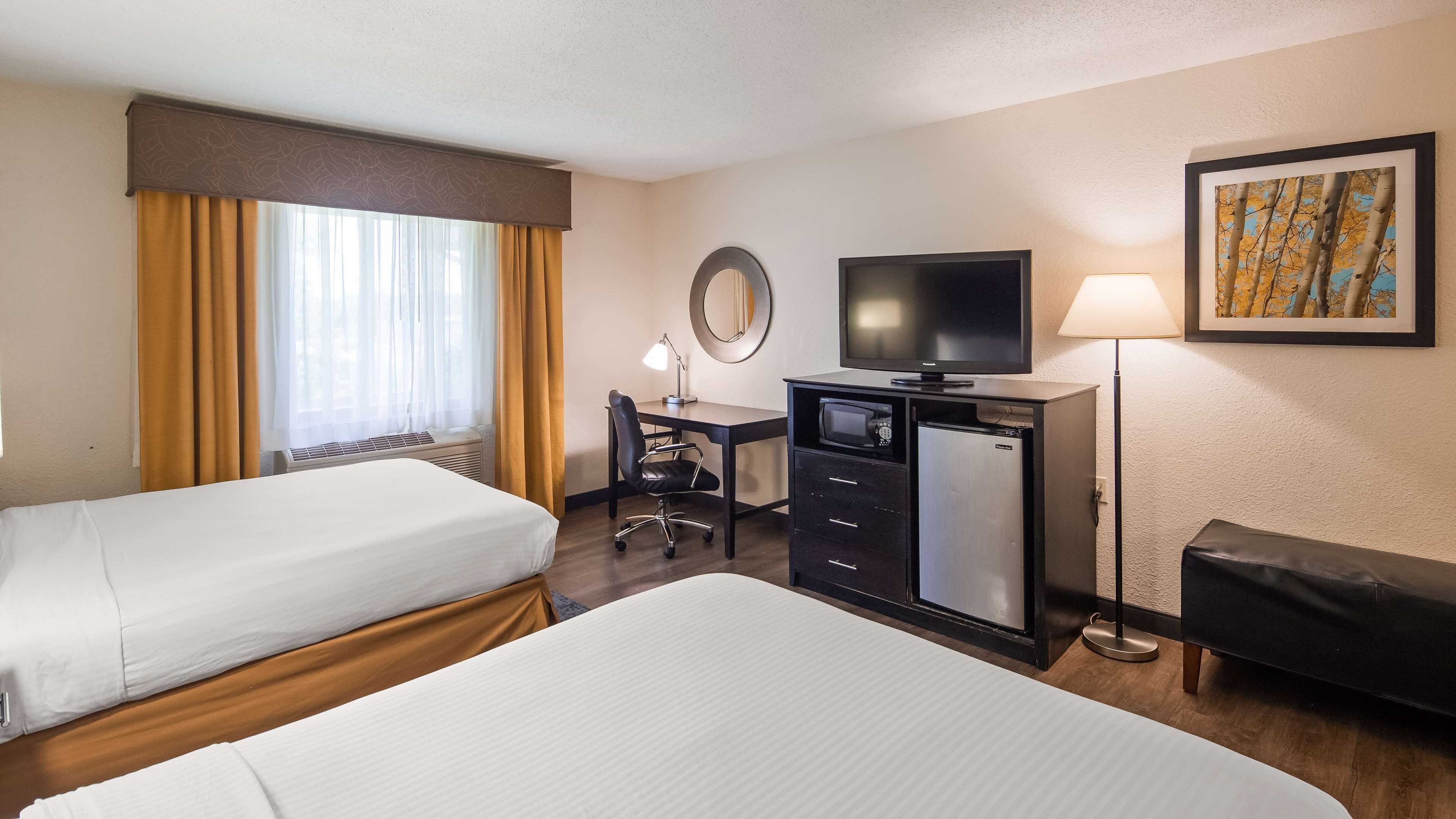 A hotel room with two beds, a desk with a computer, a television, a refrigerator, a lamp, and a painting on the wall.