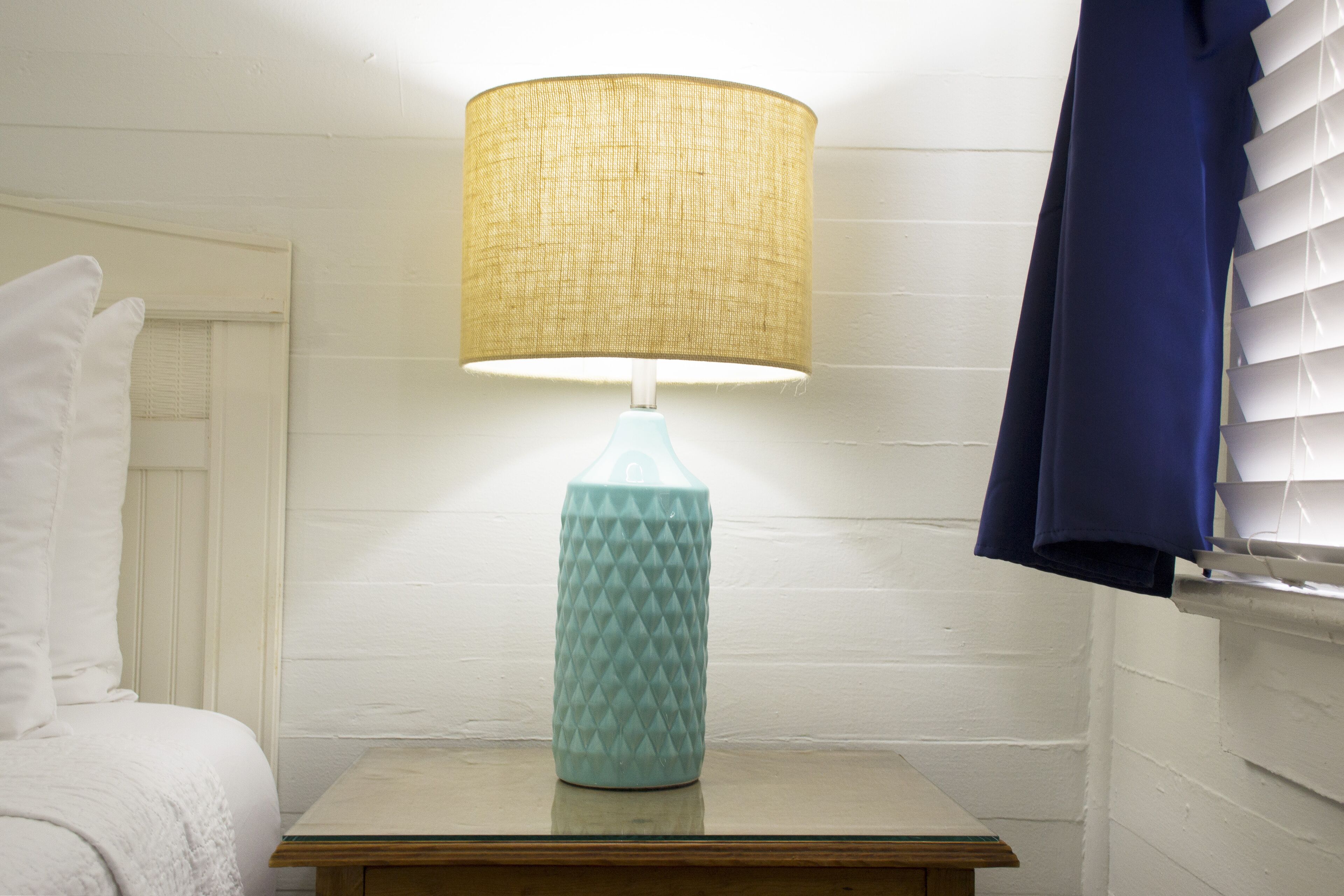 A bed with white bedding, a small table with a teal lamp, and a window with blinds.