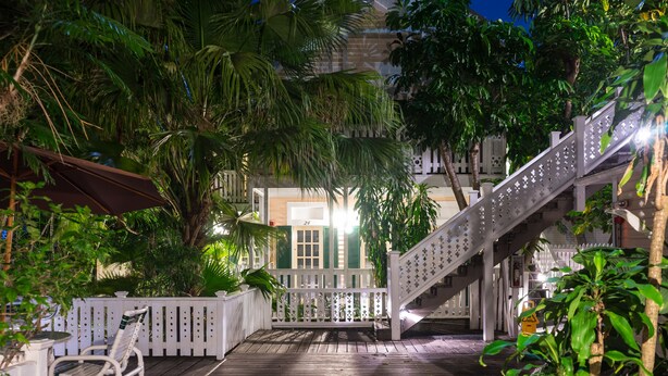 A wooden deck with white railings, surrounded by lush greenery and a staircase leading to a building. - Duval House (Key West)