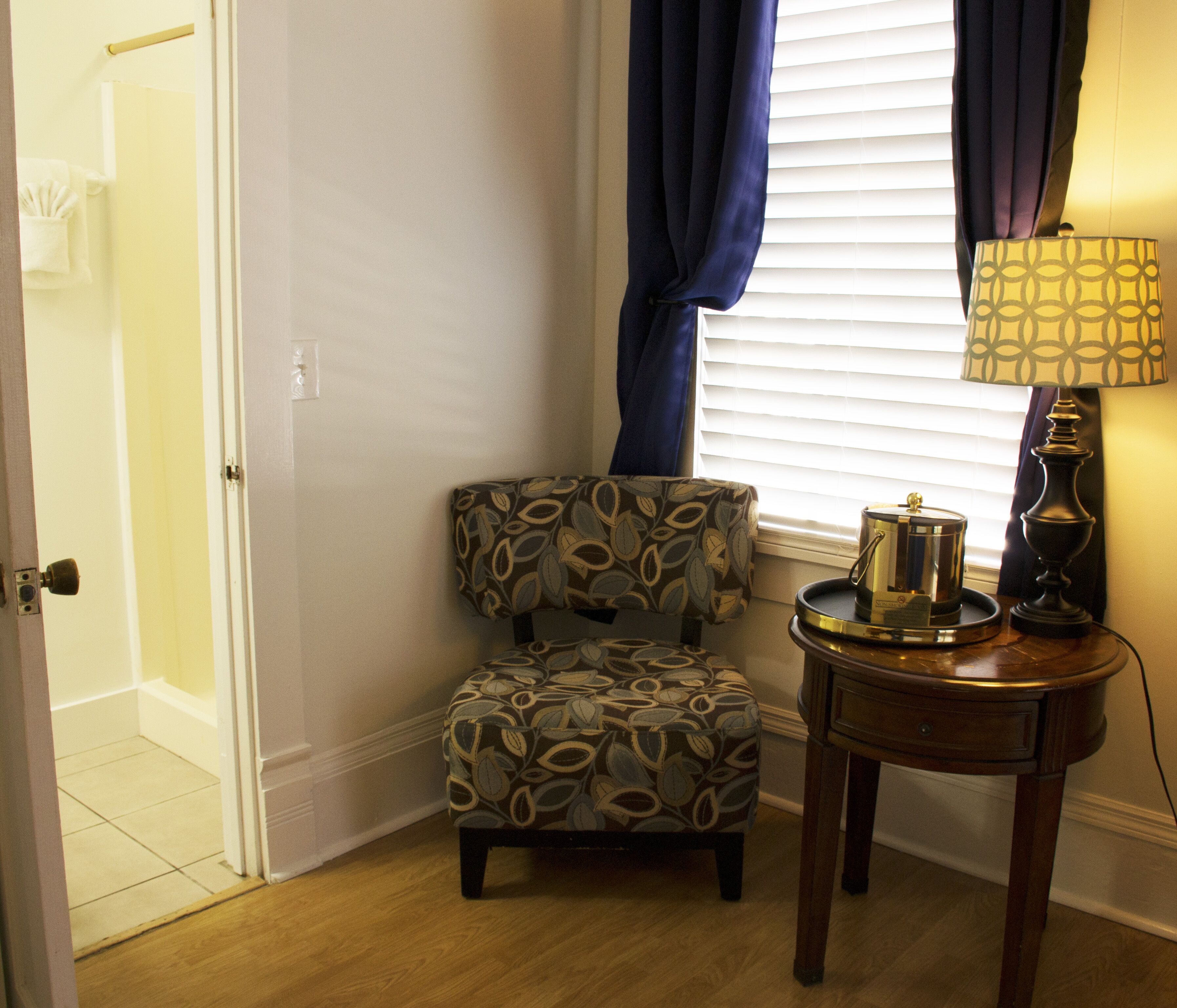A corner room with a patterned armchair, a small table with a lamp, and a window with blinds.