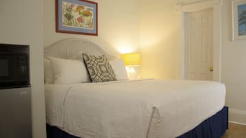 Una cama bien hecha con cubrecama blanco y almohada estampada, una lámpara de noche, una obra de arte floral enmarcada en la pared y una puerta cerrada.