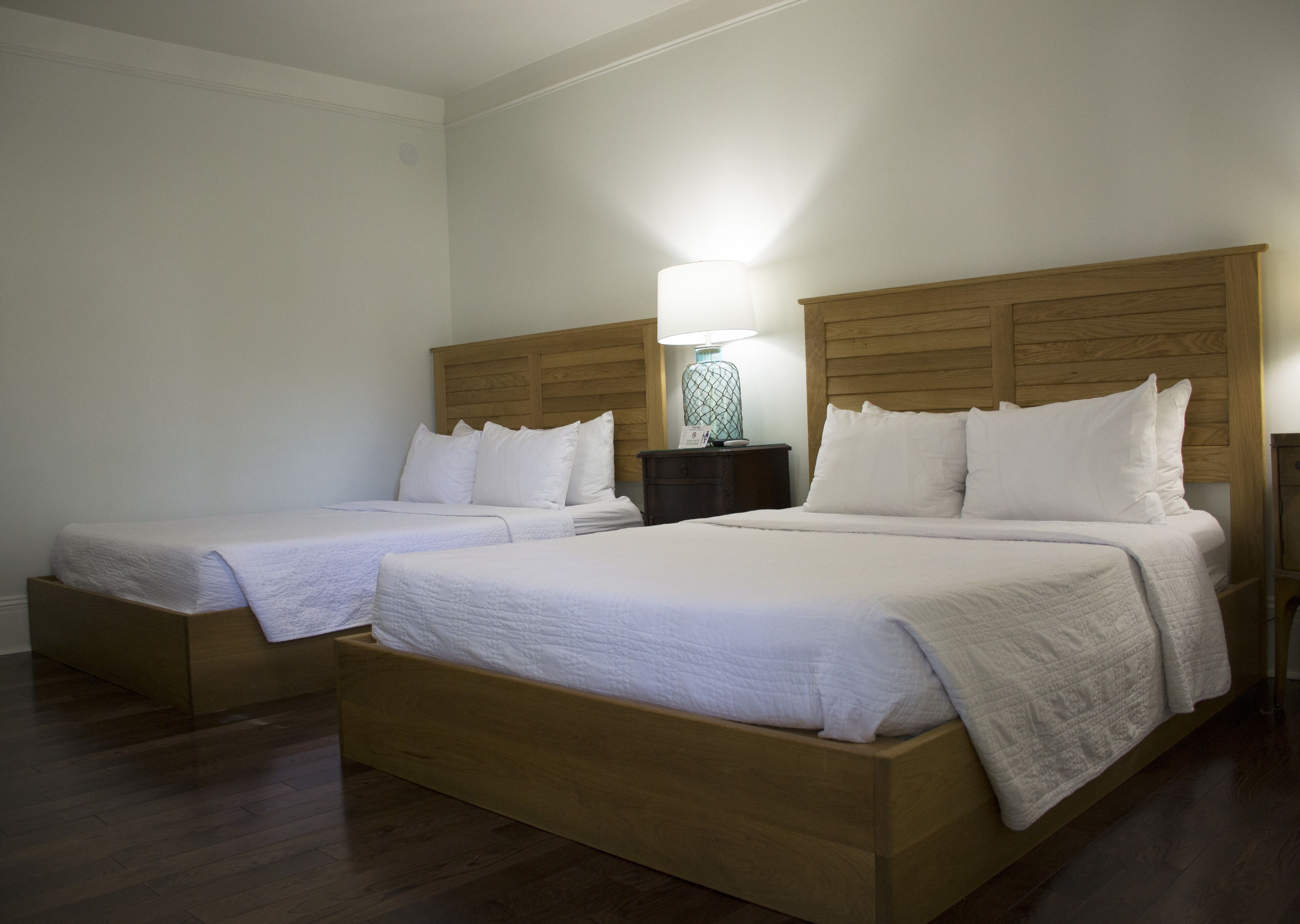 A hotel room with two single beds, wooden headboards, white bedding, a nightstand with a lamp, and a wooden floor.