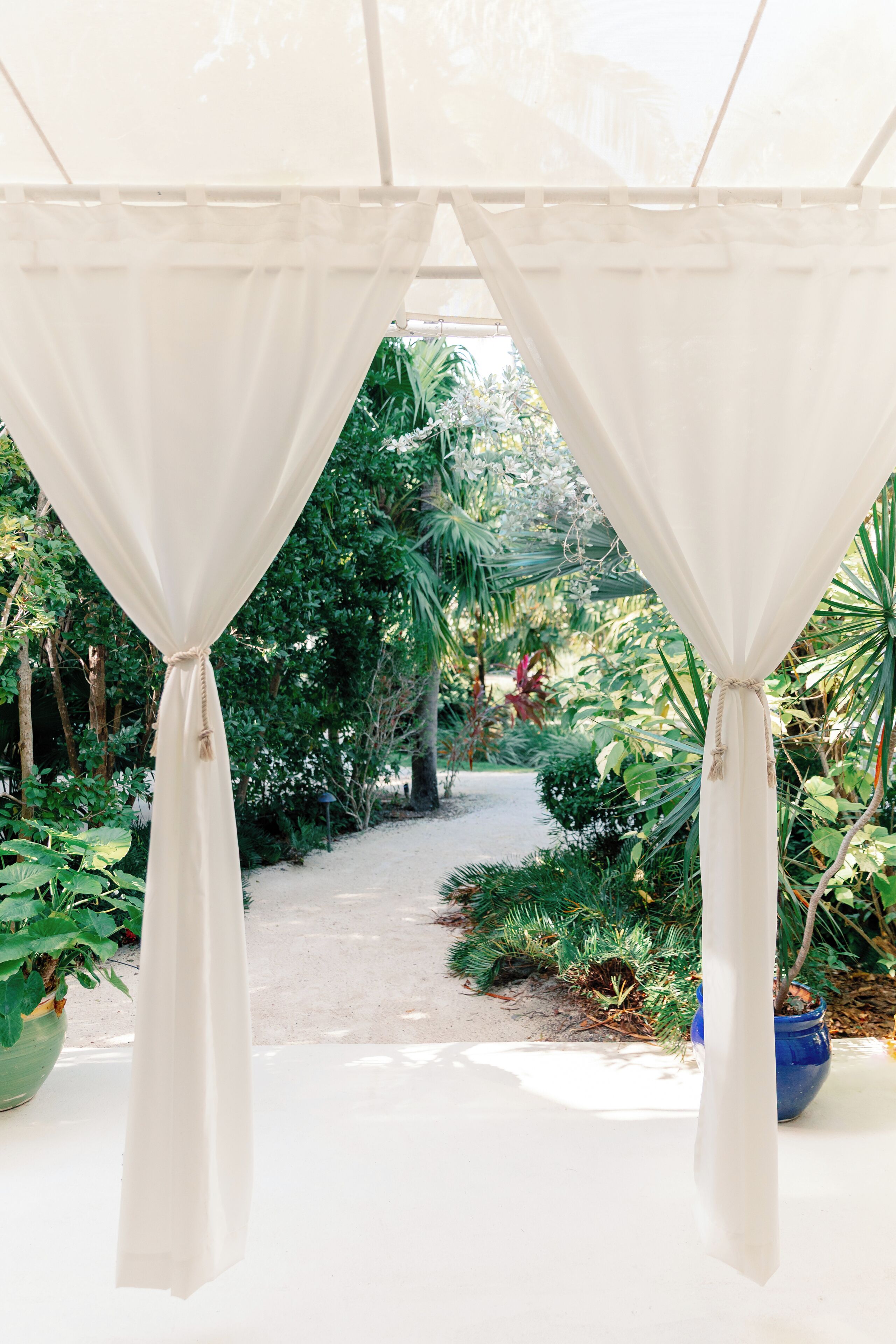 A covered pathway with white curtains, surrounded by lush greenery and potted plants.