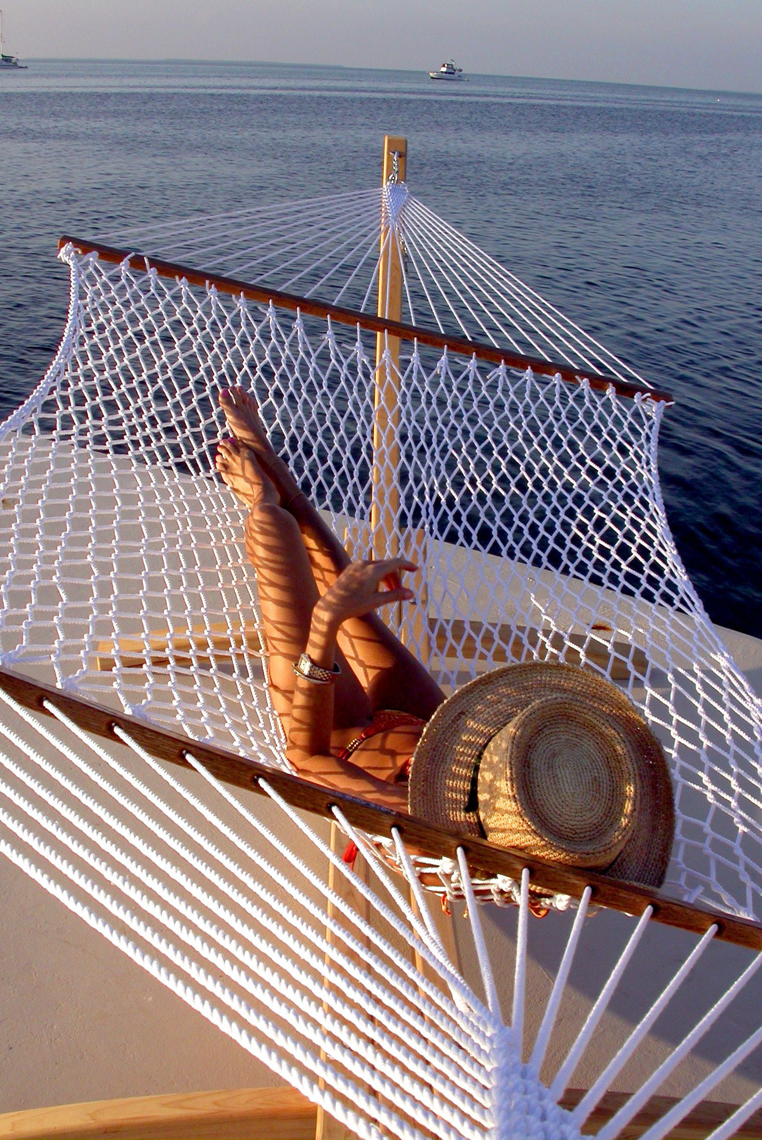 Un hamac dans lequel est allongé une personne, un chapeau de paille et une vue sur la mer avec des bateaux à l’horizon.