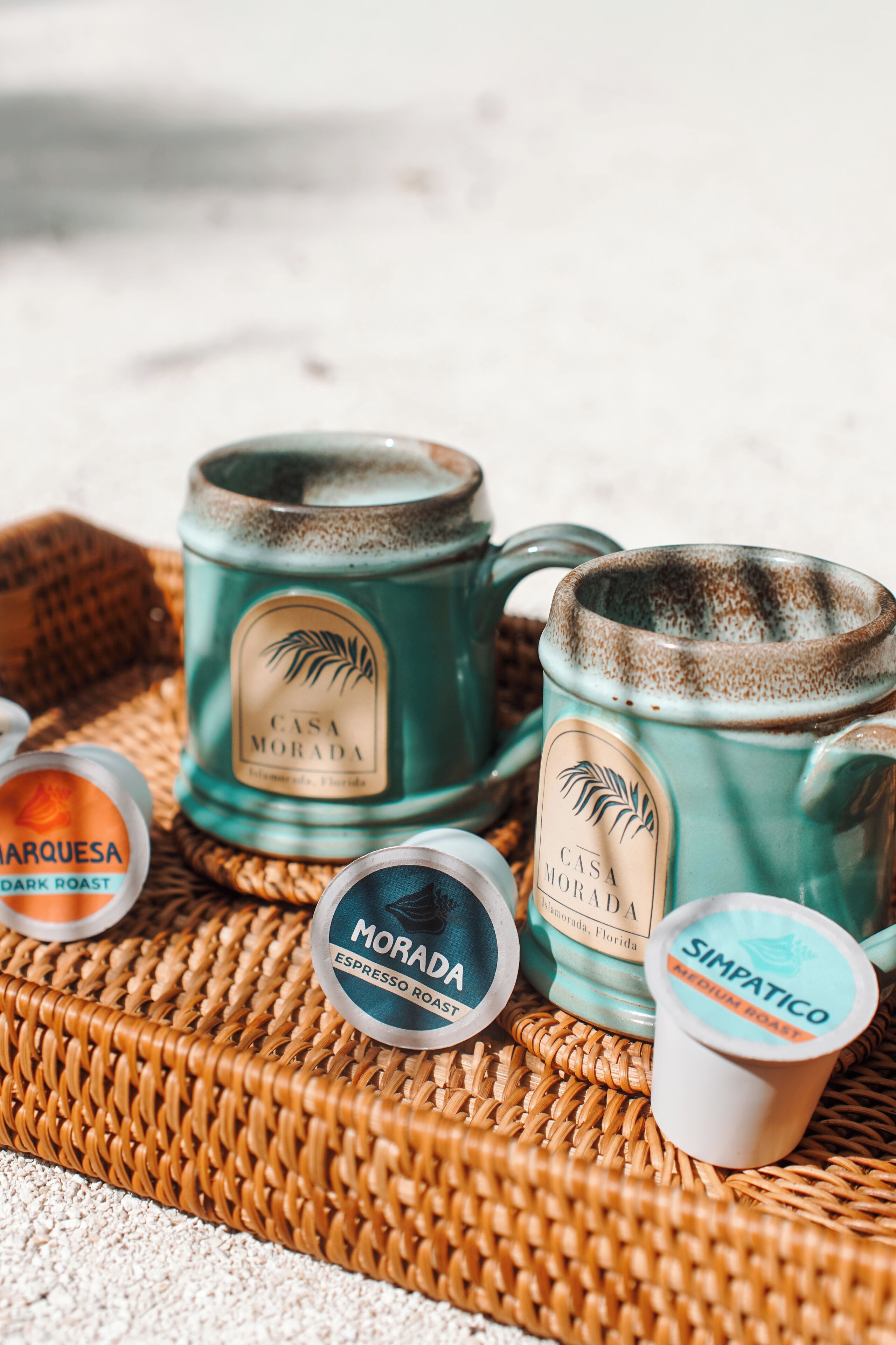 Two teal ceramic mugs with labels, a small jar of dark roast coffee, and a cup of medium roast coffee on a woven mat.
