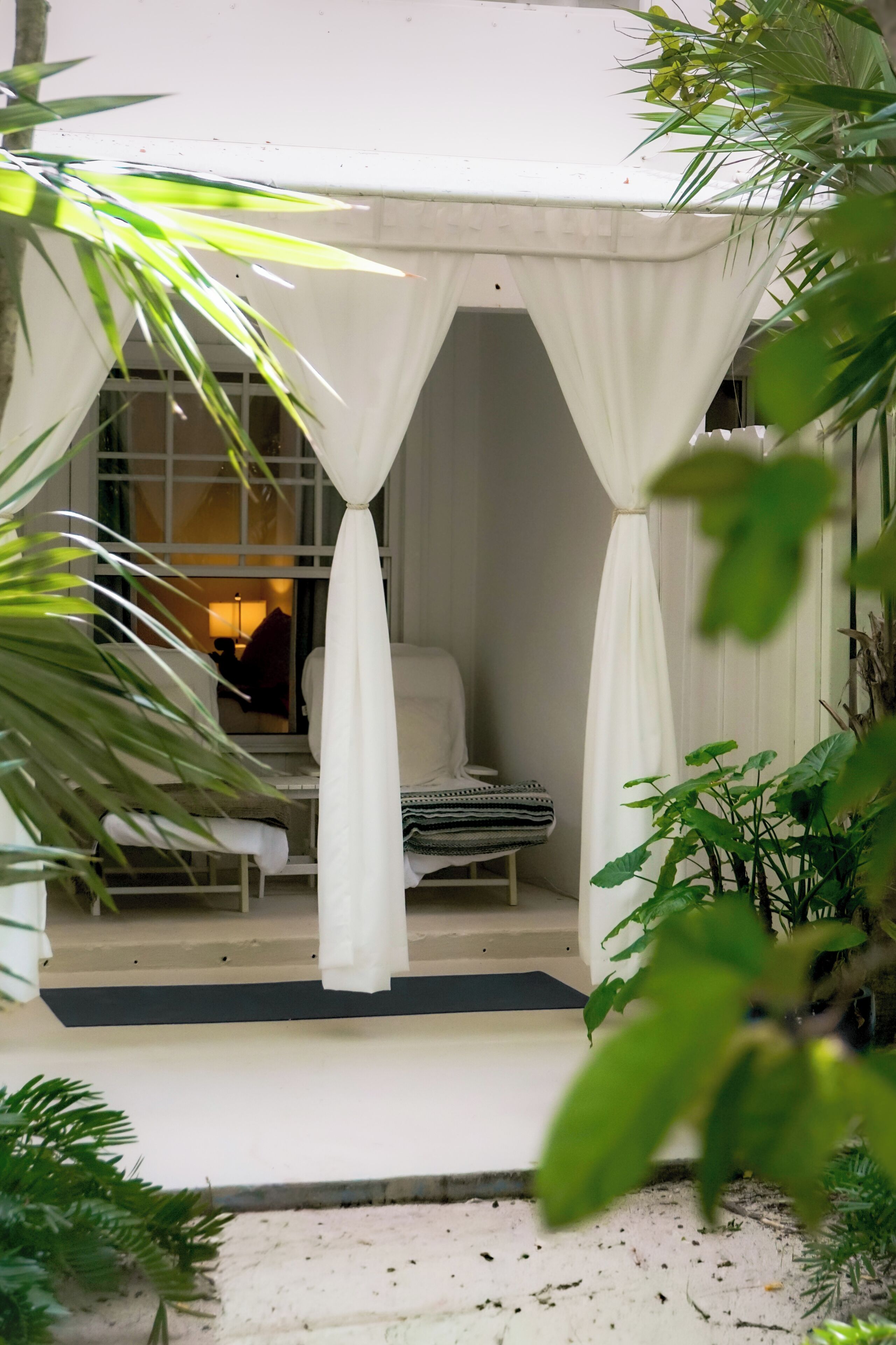 A covered outdoor seating area with a white chair and a patterned bench, surrounded by greenery.