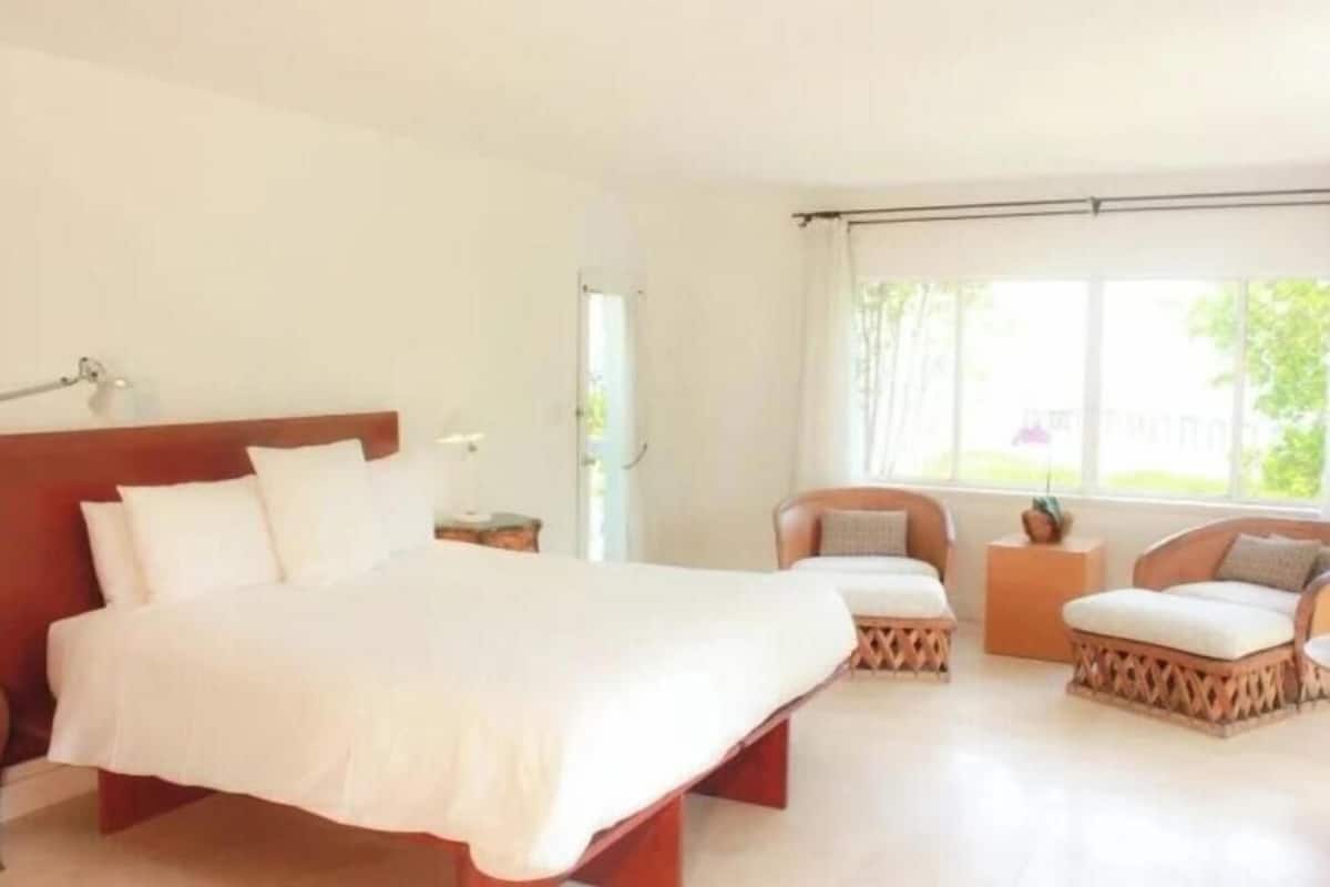 Quarto de hotel com uma cama grande, duas poltronas, uma mesinha e uma janela com vista para o verde.