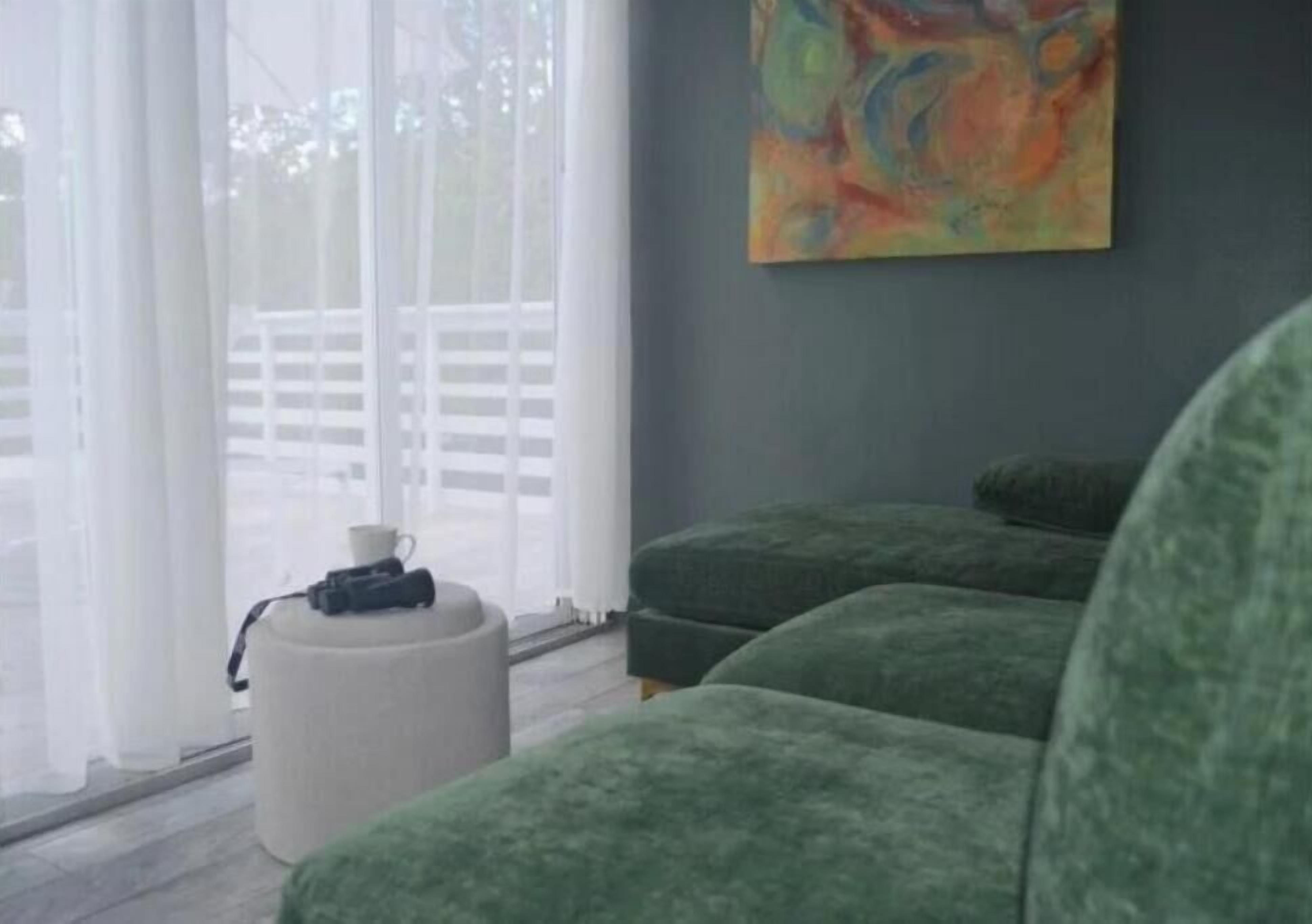 A room with a large window, a green sofa set, a white ottoman, and a colorful abstract painting on the wall.