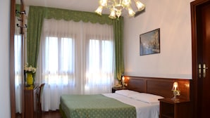 A hotel room with a bed, a nightstand with a lamp, a desk with a chair, a window with curtains, and a painting on the wall. - Hotel Villa Cipro (Venice)