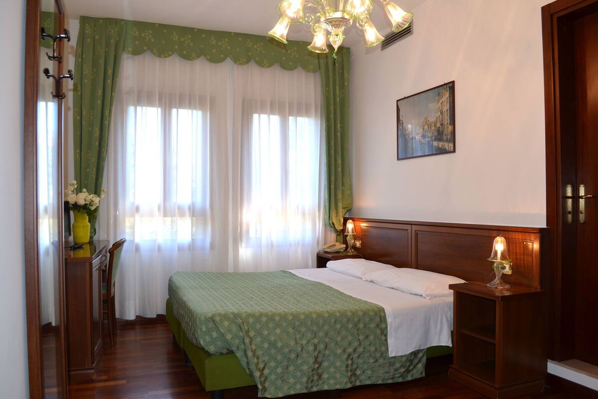 Una habitación de hotel con cama, mesita de noche con lámpara, escritorio con silla, ventana con cortinas y un cuadro en la pared.