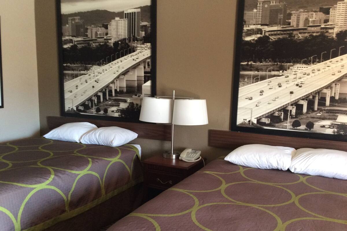 A hotel room with two beds, a nightstand with a lamp, and a cityscape view.