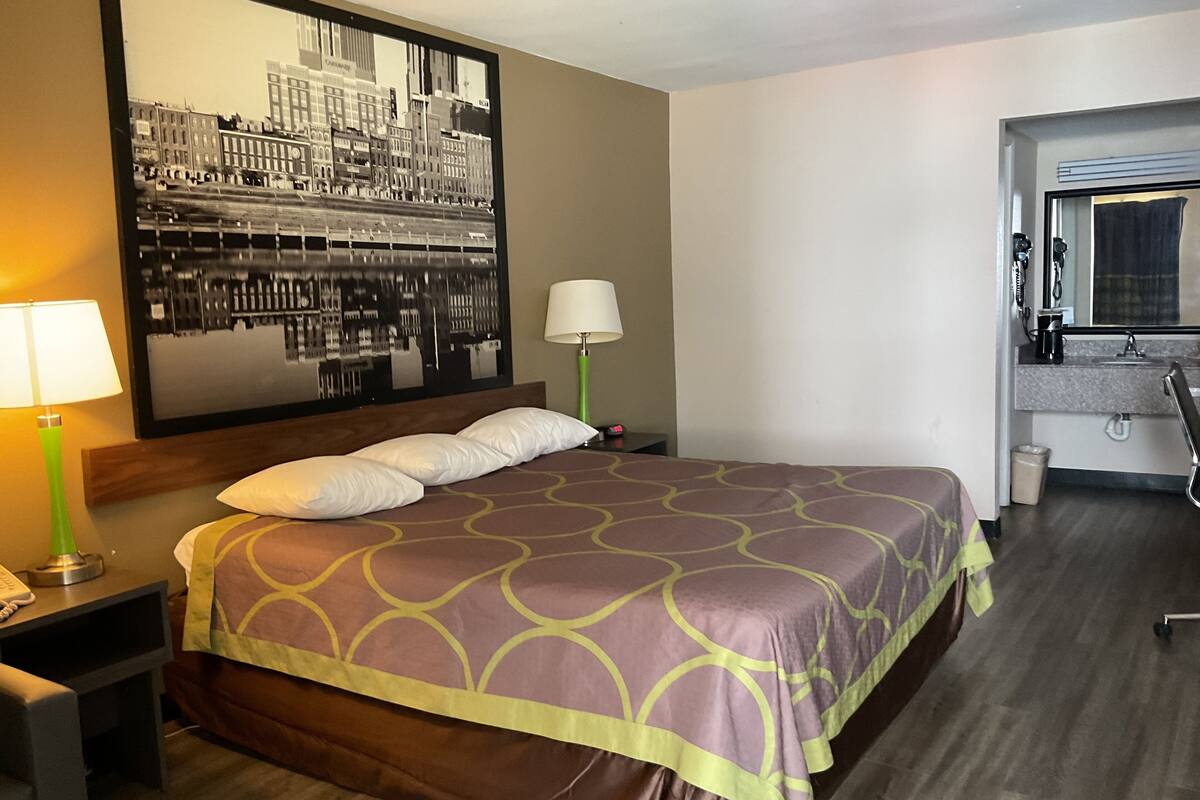 A hotel room with a large bed, a nightstand with a lamp, a wall-mounted mirror, and a view of the cityscape.
