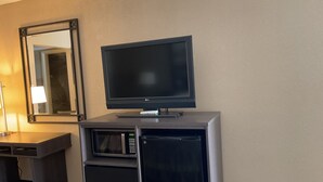 Hotellihuone, jossa on jalustalla oleva taulutelevisio, mikroaaltouuni ja pieni työpöytä, jossa on lamppu.
