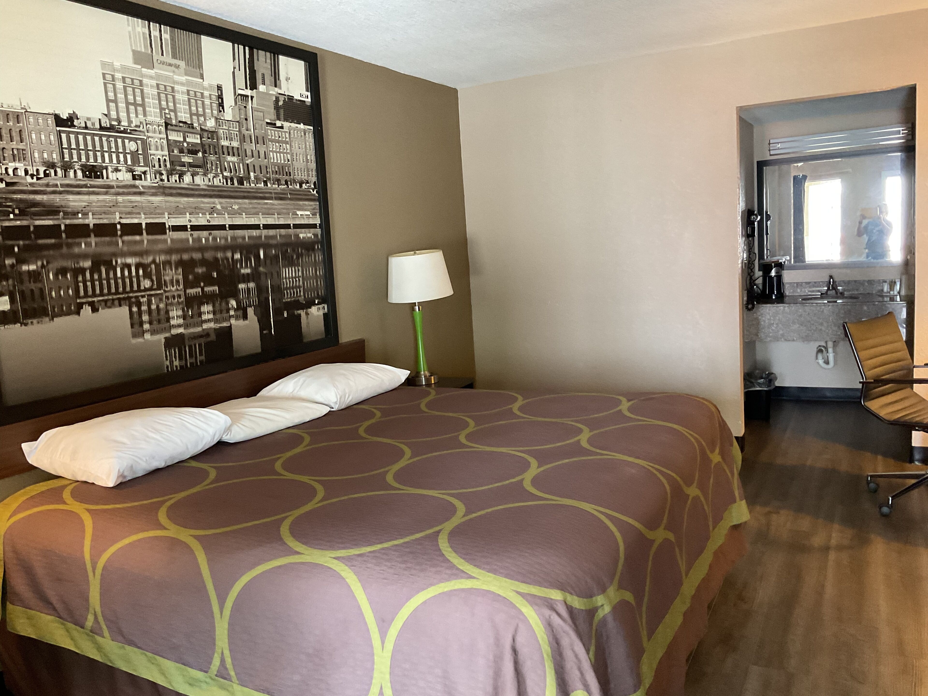 Une chambre d’hôtel avec un grand lit, un miroir reflétant le paysage urbain, une lampe de sol verte et une kitchenette.