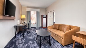 A hotel room with a sofa, a small table, a desk with a chair, a TV mounted on the wall, a lamp, and a microwave. - Fairfield Inn by Marriott Toronto Oakville (Oakville)