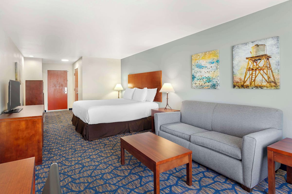 A hotel room with a bed, a grey sofa, a wooden coffee table, a TV, and two paintings on the wall.