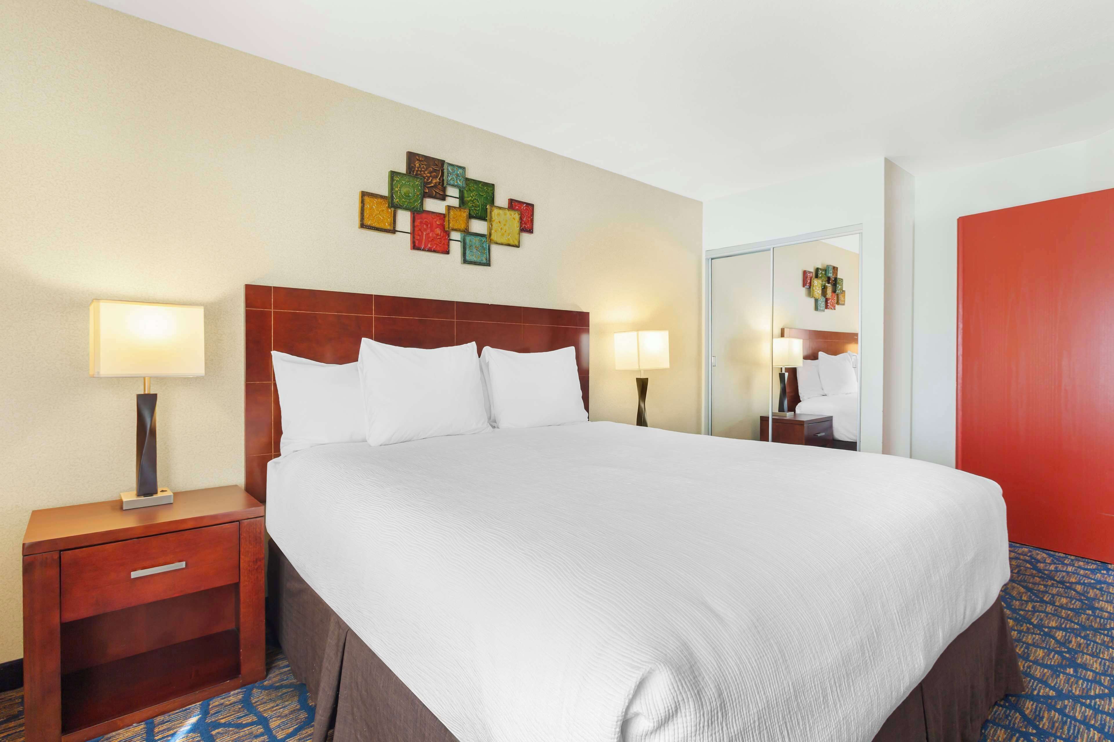 Una habitación de hotel con una cama grande, dos lámparas de noche, una mesita de noche de madera, un espejo y una decoración de pared colorida.