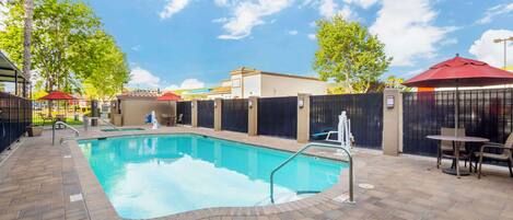 A swimming pool with a tiled deck, surrounded by a fence and shaded by red umbrellas.