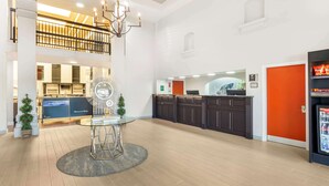 A spacious lobby with a reception desk, a dining area with a round table, and a kitchen area with dark cabinets and an orange accent wall. - Best Western Plus Villa Del Lago Inn Patterson (Patterson)