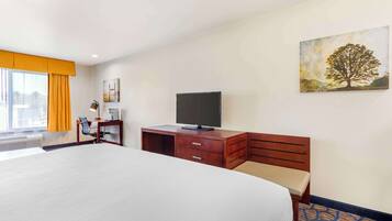 A hotel room with a large bed, a desk with a flat-screen TV, a chair, a small table with a lamp, and a painting of a tree on the wall.