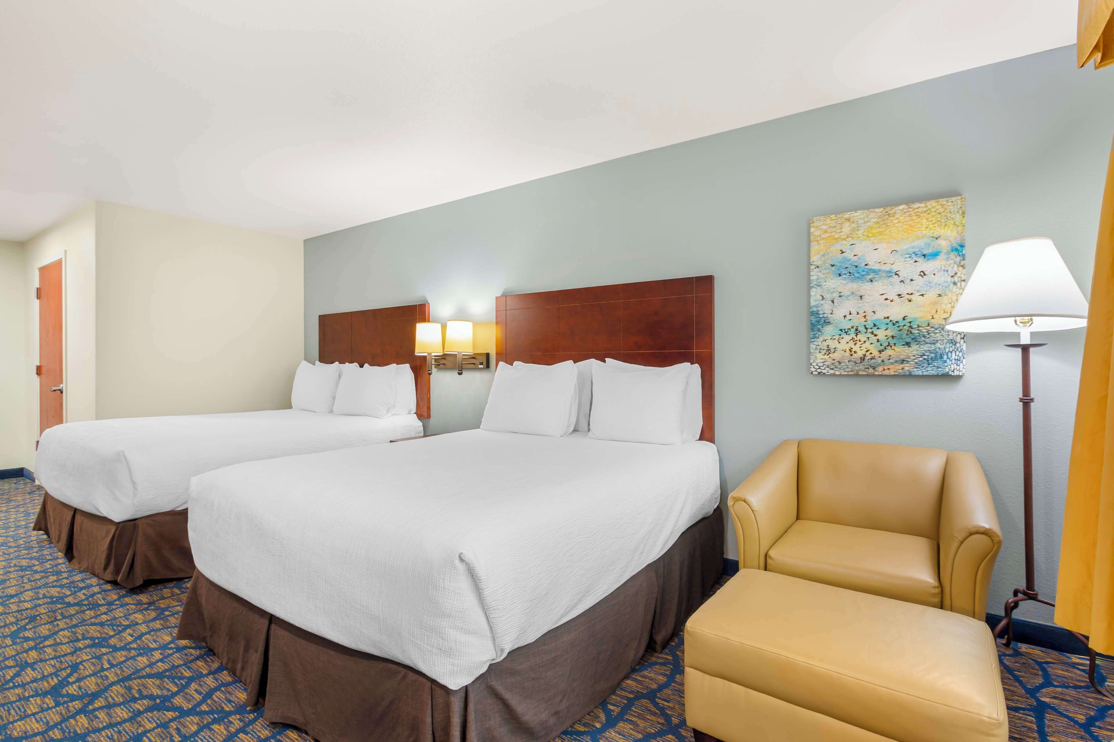 A hotel room with two beds, a chair, a lamp, and a painting on the wall.