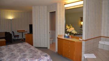 Une chambre d’hôtel comprenant un lit, un bureau, une chaise, une salle de bain avec un lavabo et un miroir, ainsi qu’un espace douche.