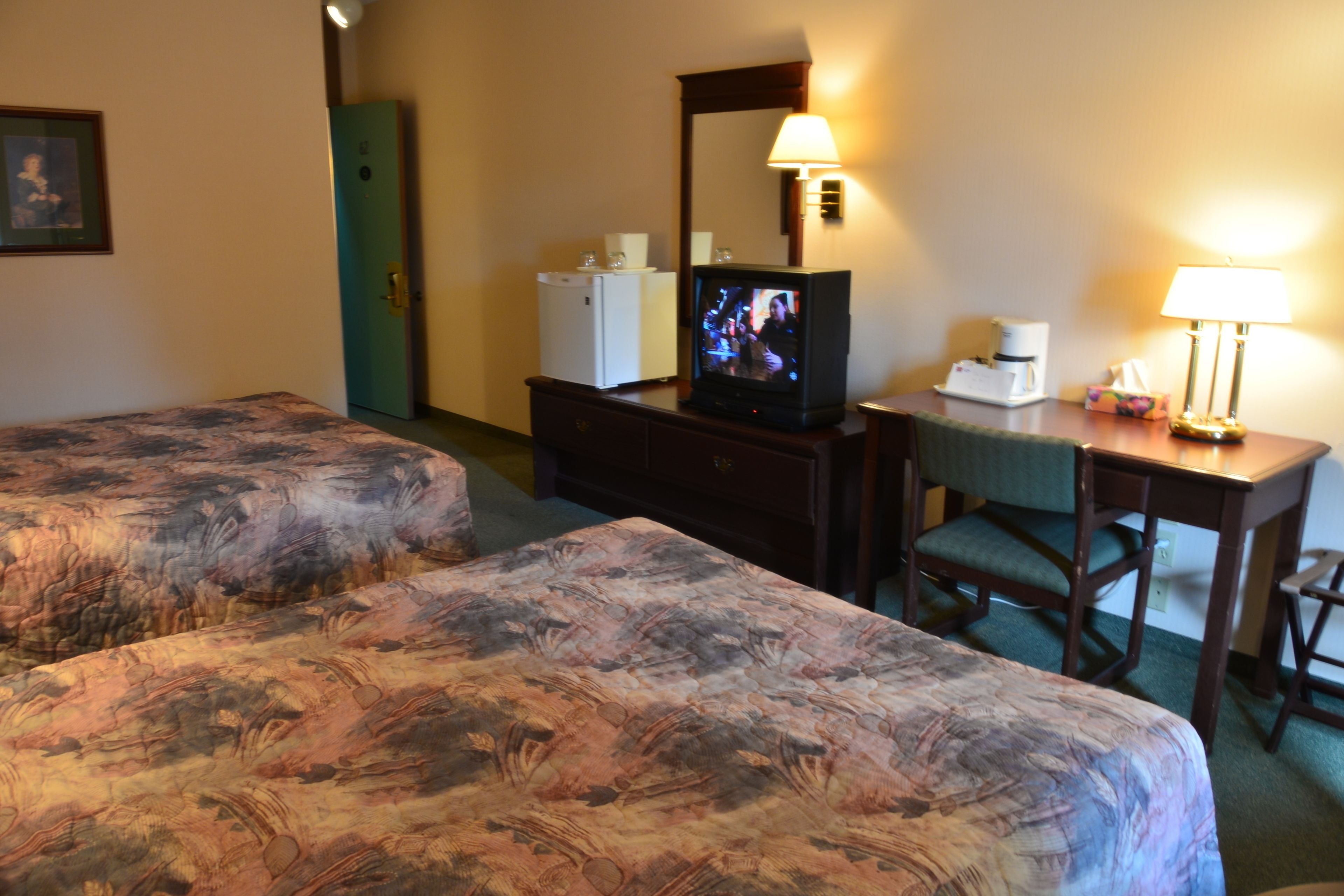 A hotel room with two beds, a desk, a television, a mirror, a lamp, and a refrigerator.