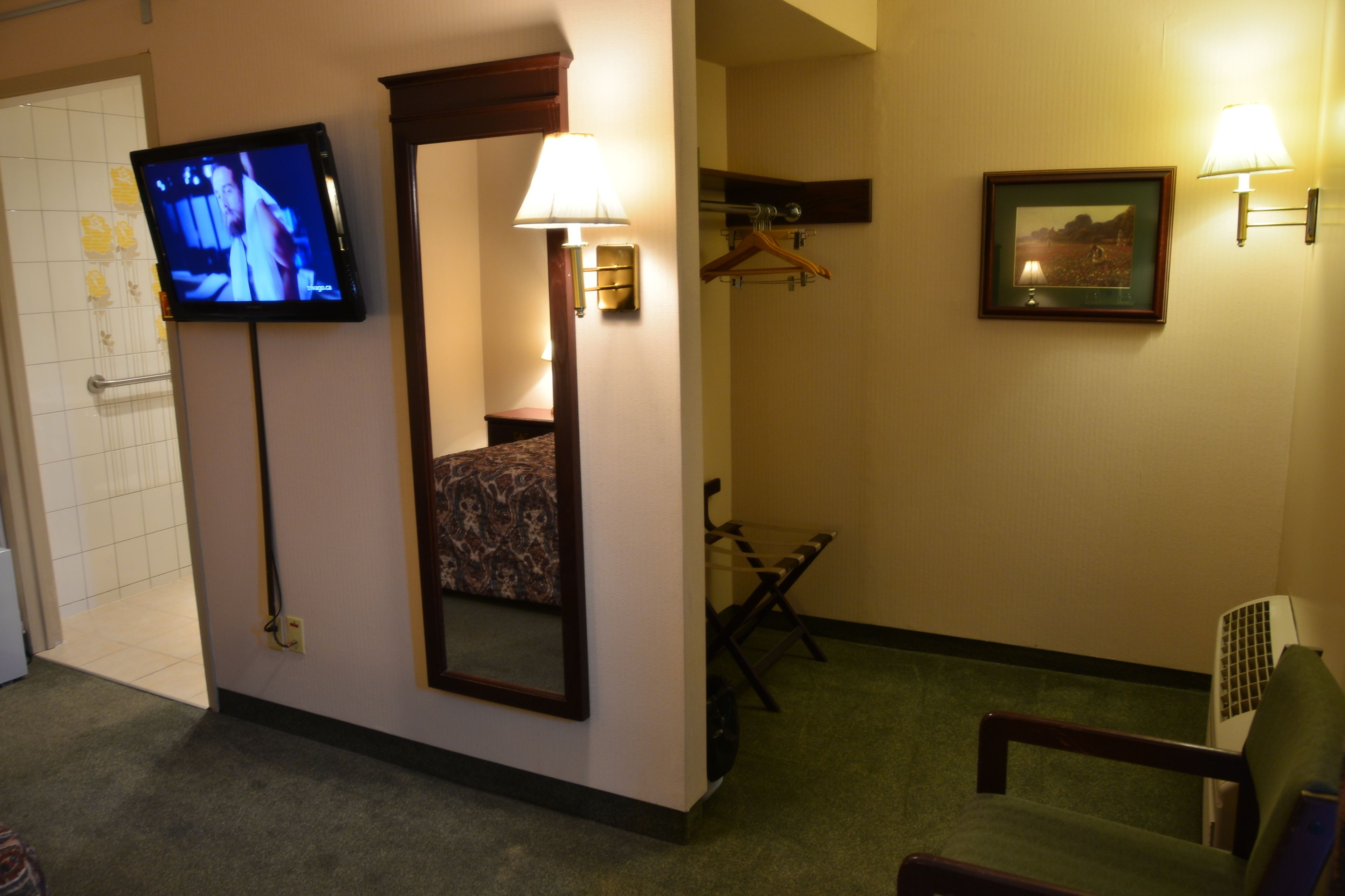 A hotel room with a television, a mirror, a bed, a chair, a hanging coat rack, and a picture on the wall.
