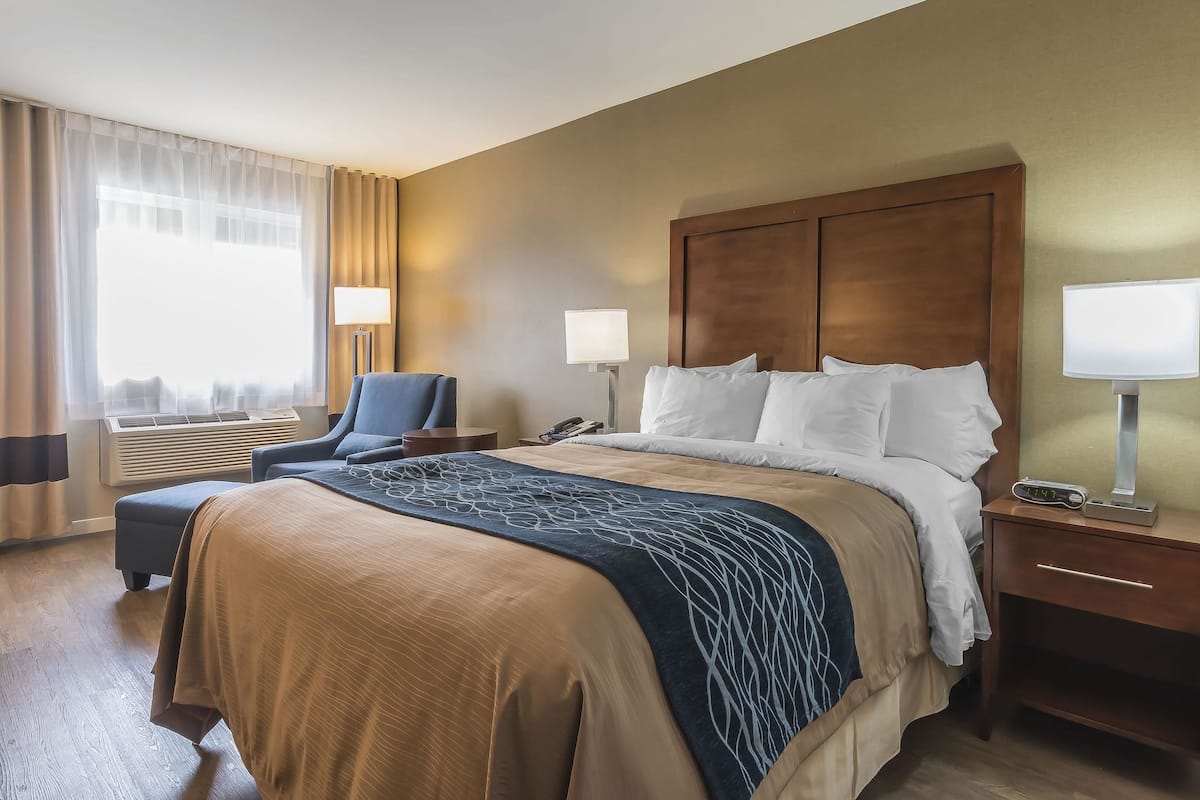 Una habitación de hotel con una cama grande, una mesita de noche con lámpara, una silla y un ventanal con cortinas.