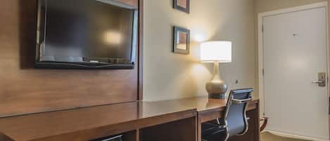 Habitación de hotel con escritorio de madera, televisor, lámpara, silla y minibar.
