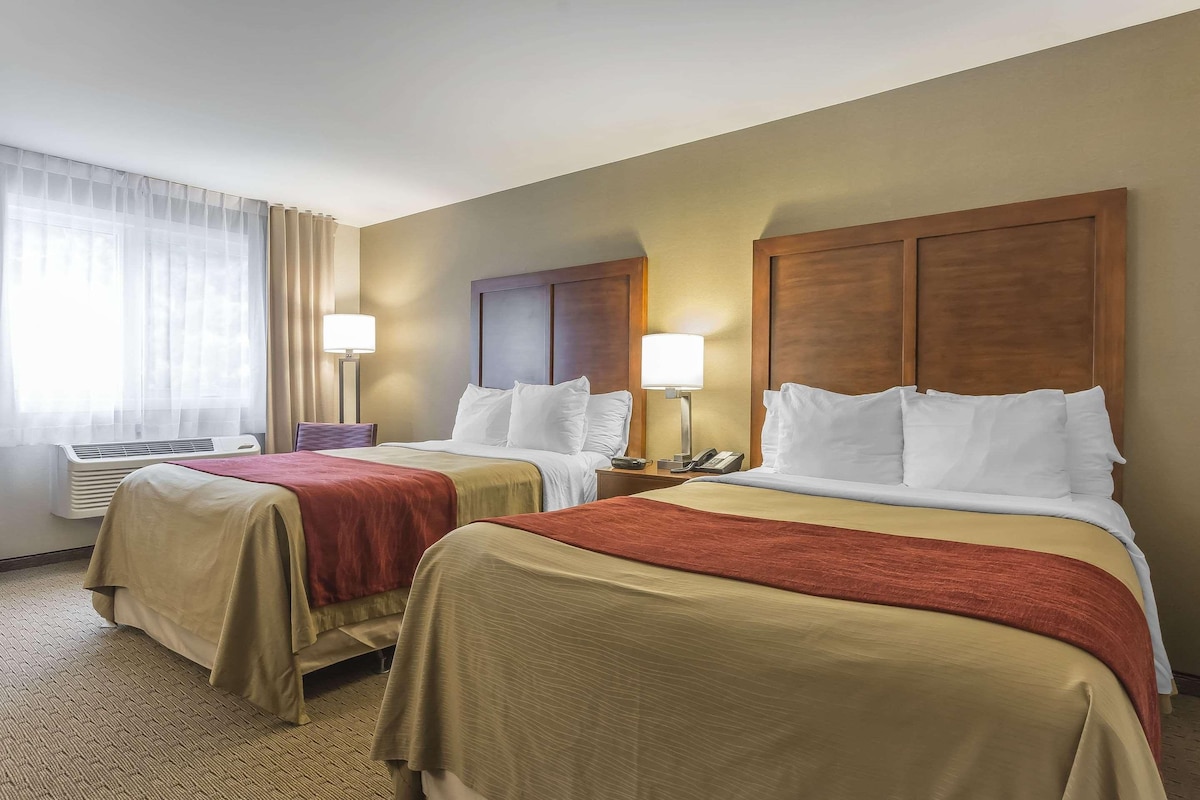 A hotel room with two beds, each with a wooden headboard, beige bedding, and a red throw blanket. There is a window with sheer curtains, a bedside table with a lamp, and an air conditioning unit.