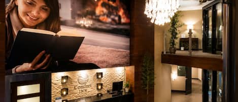 A hotel reception area with a large poster, a reception desk, a chandelier, and a staircase.