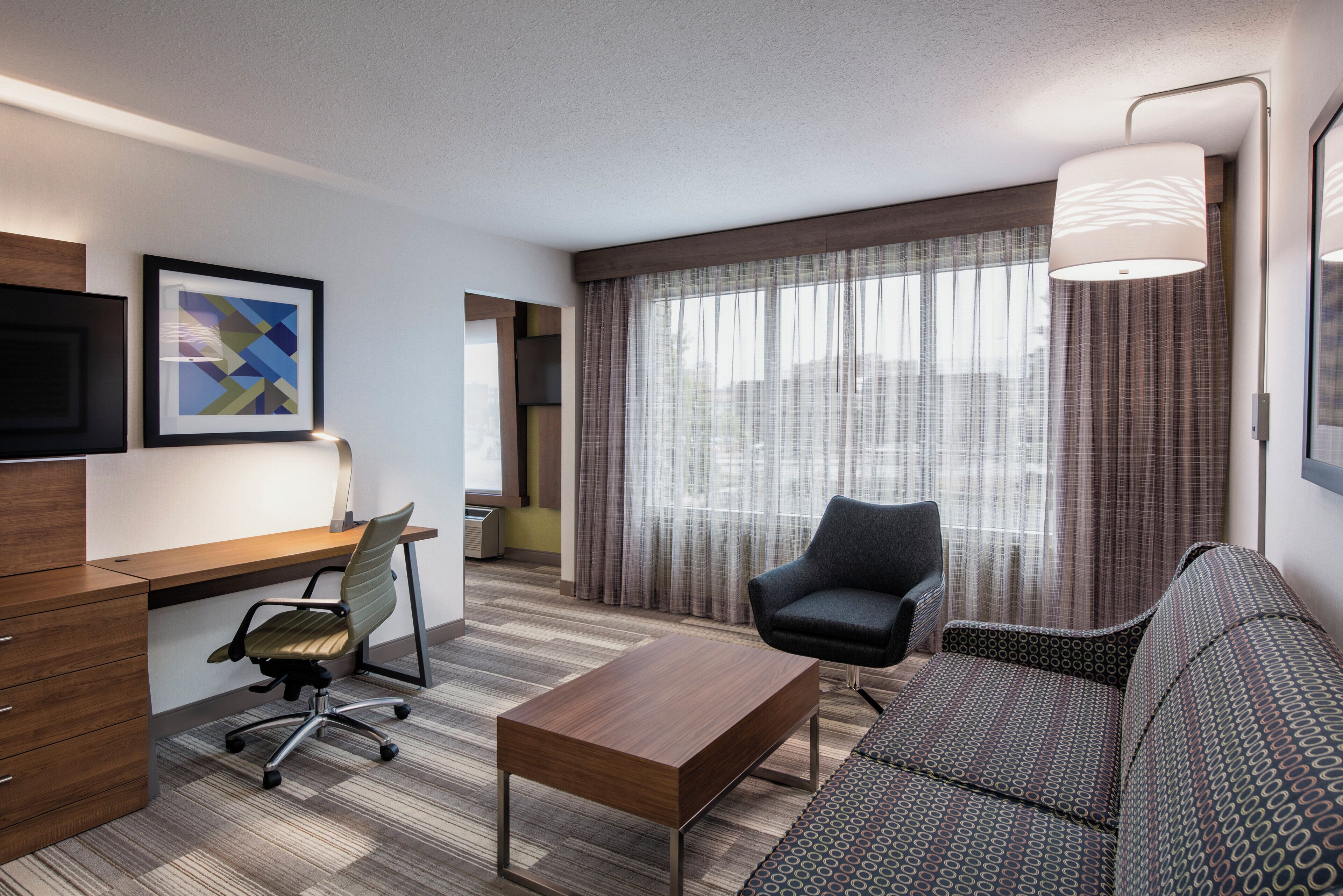 Una habitación de hotel con escritorio, silla de oficina, sofá y un ventanal con cortinas.