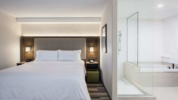 A modern hotel room with a large bed, bedside tables, a nightstand, a green ottoman, a glass shower enclosure, and a white bathroom with a bathtub. - Holiday Inn Express & Suites Saskatoon Centre by IHG (Saskatoon)