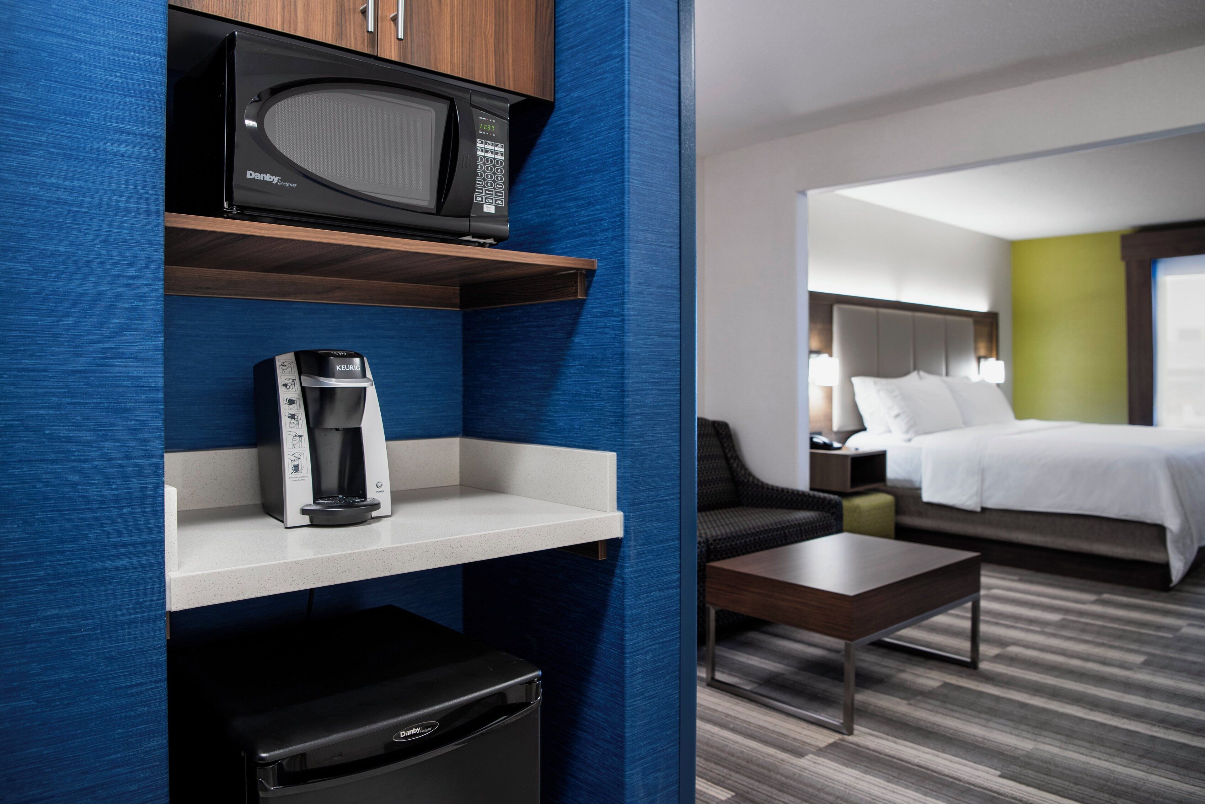 Une chambre d’hôtel équipée d’un four à micro-ondes, d’une machine à café et d’un petit réfrigérateur posé sur une étagère. On y trouve un lit avec un mur d’accent vert, une table de chevet et une chaise.