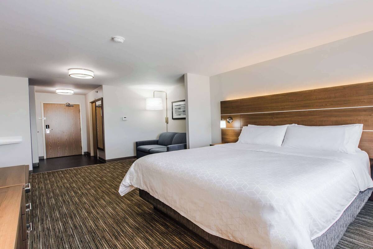 A hotel room with a large bed, a wooden headboard, a nightstand, a seating area with a chair, and a carpeted floor.