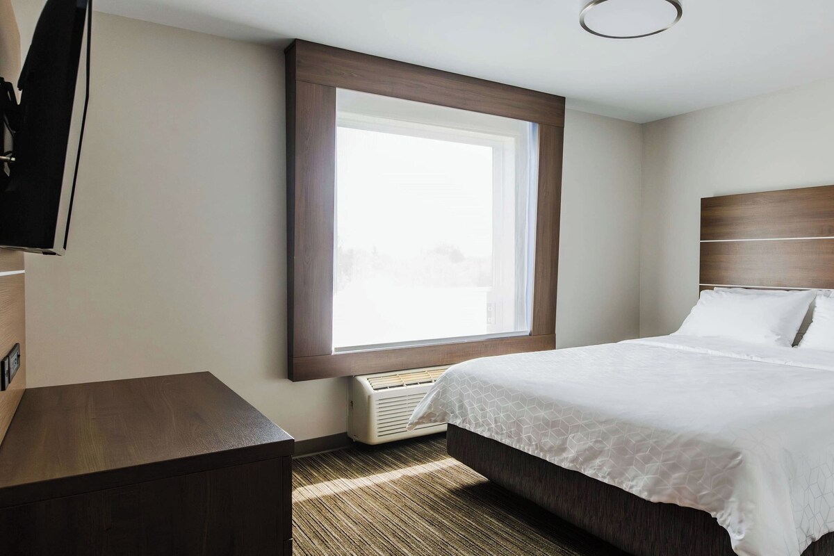 A hotel room with a large window, a bed with white linens, a wooden headboard, and a small bedside table.