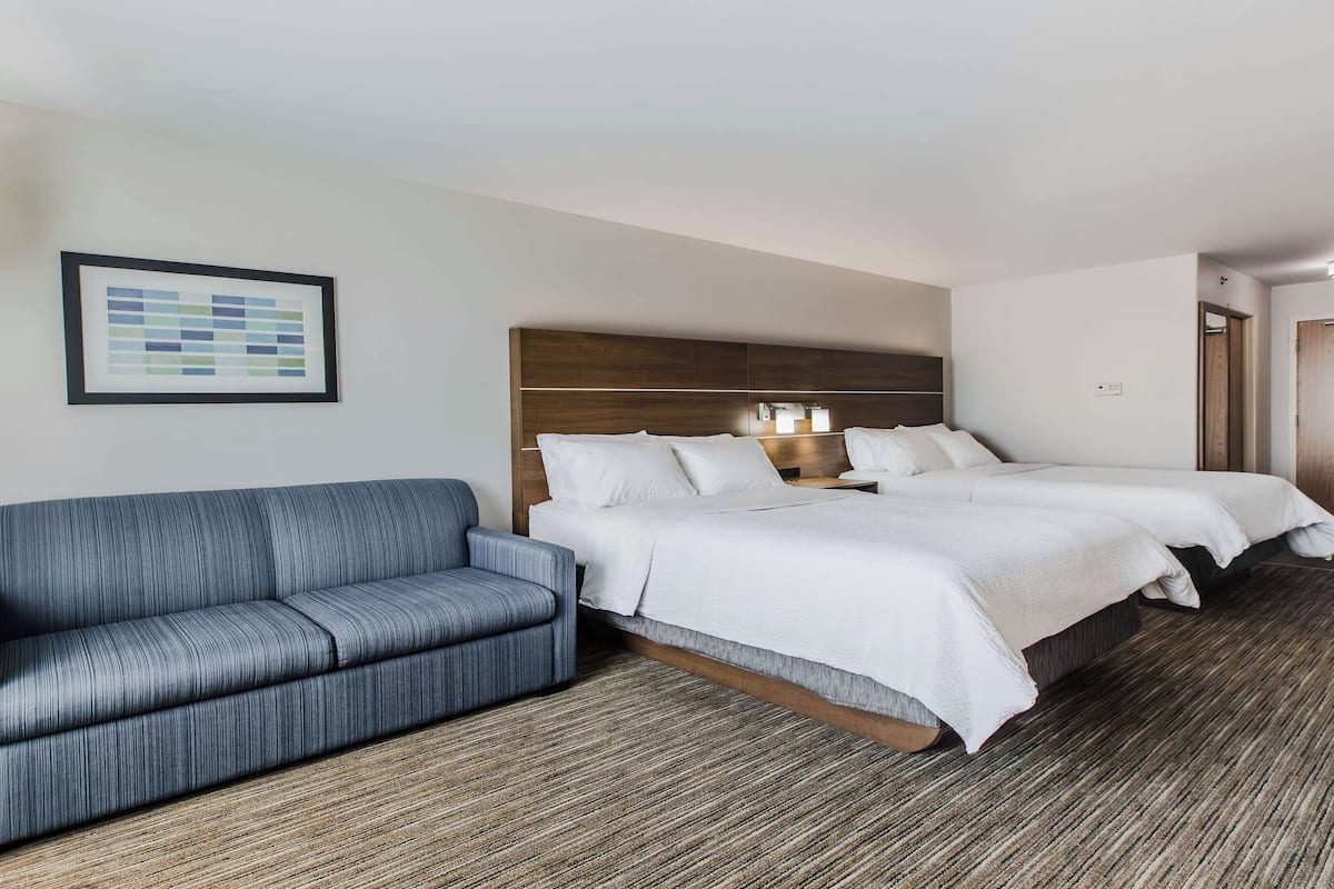 A hotel room with a sofa, two beds, a framed picture, and a lamp.