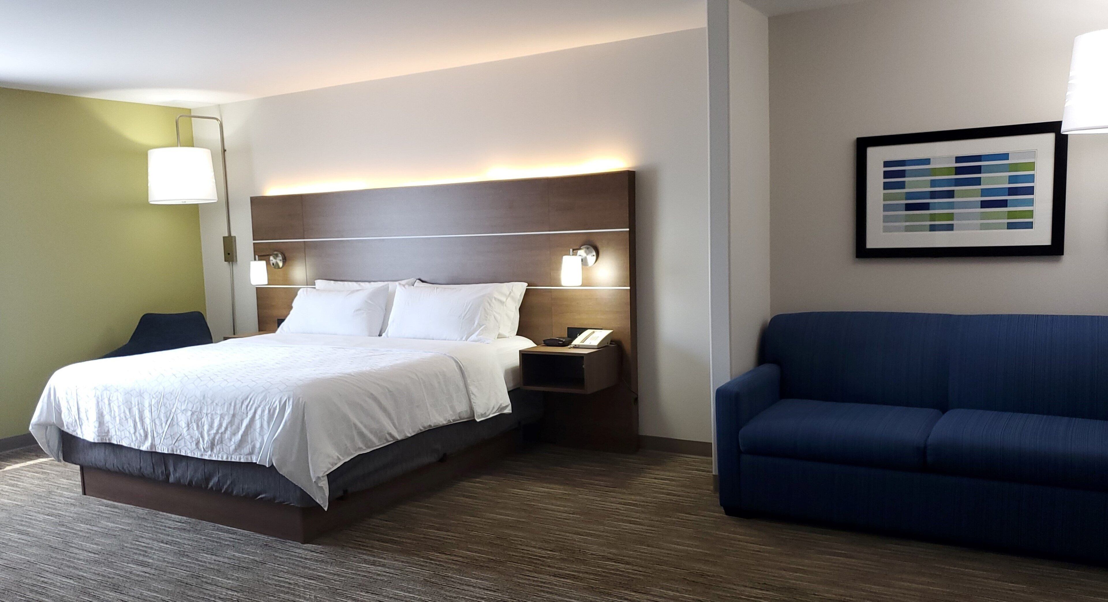 Una habitación de hotel con una cama, una mesita de noche, un sofá azul, una lámpara de pared y un cuadro enmarcado.