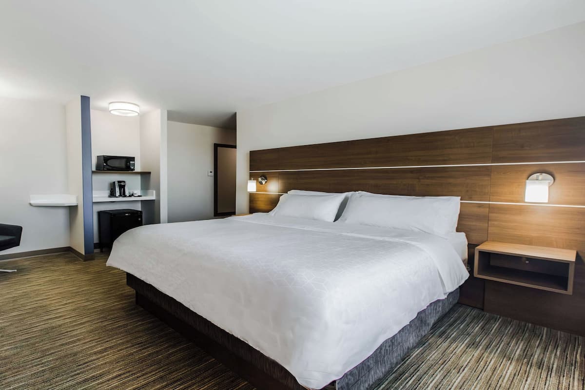 A hotel room with a large bed, a desk, a chair, a microwave, a coffee maker, and a nightstand with a lamp.