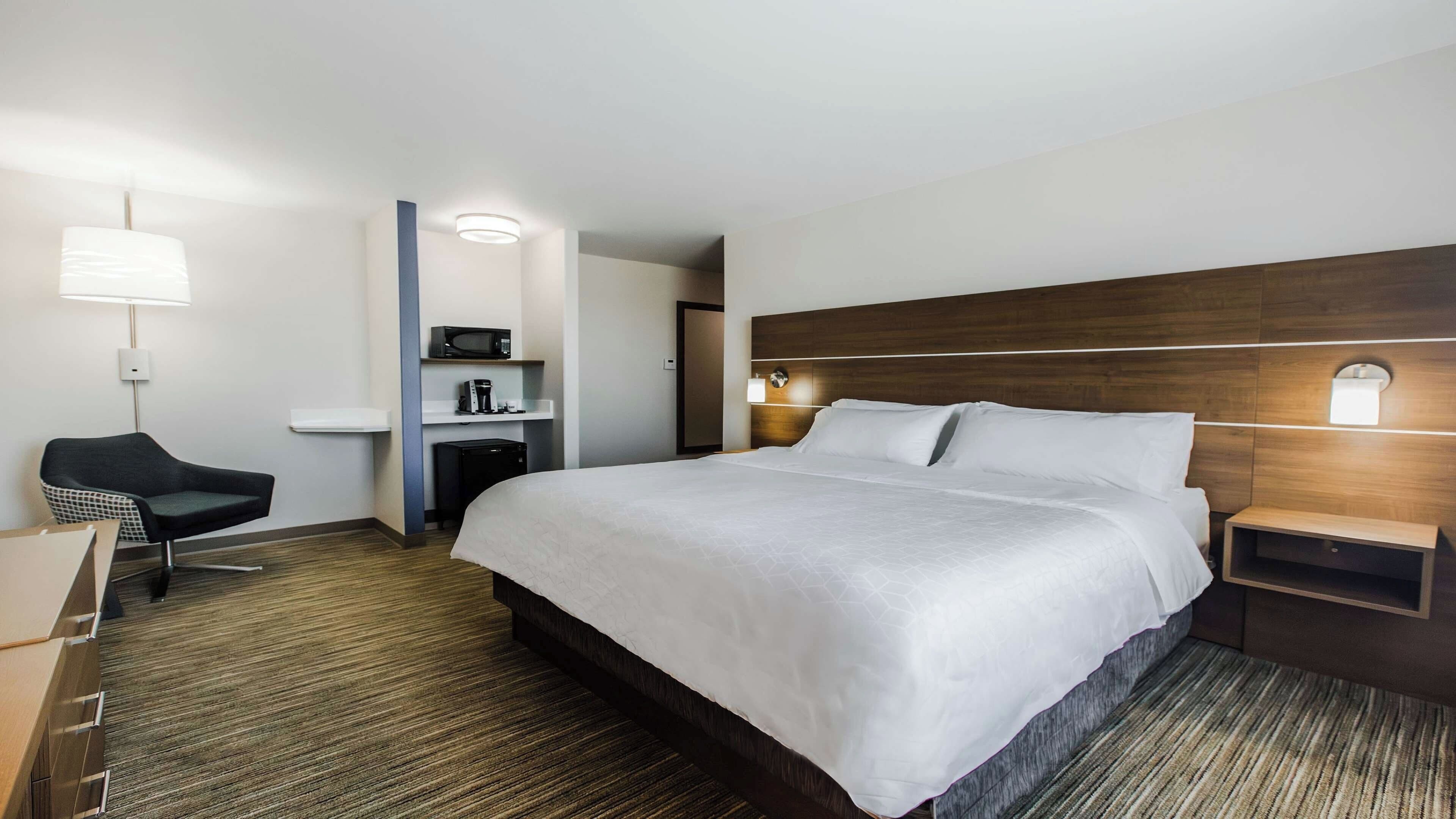 Habitación de hotel con una cama grande, un escritorio, una silla, microondas, cafetera y una mesita de noche con lámpara.