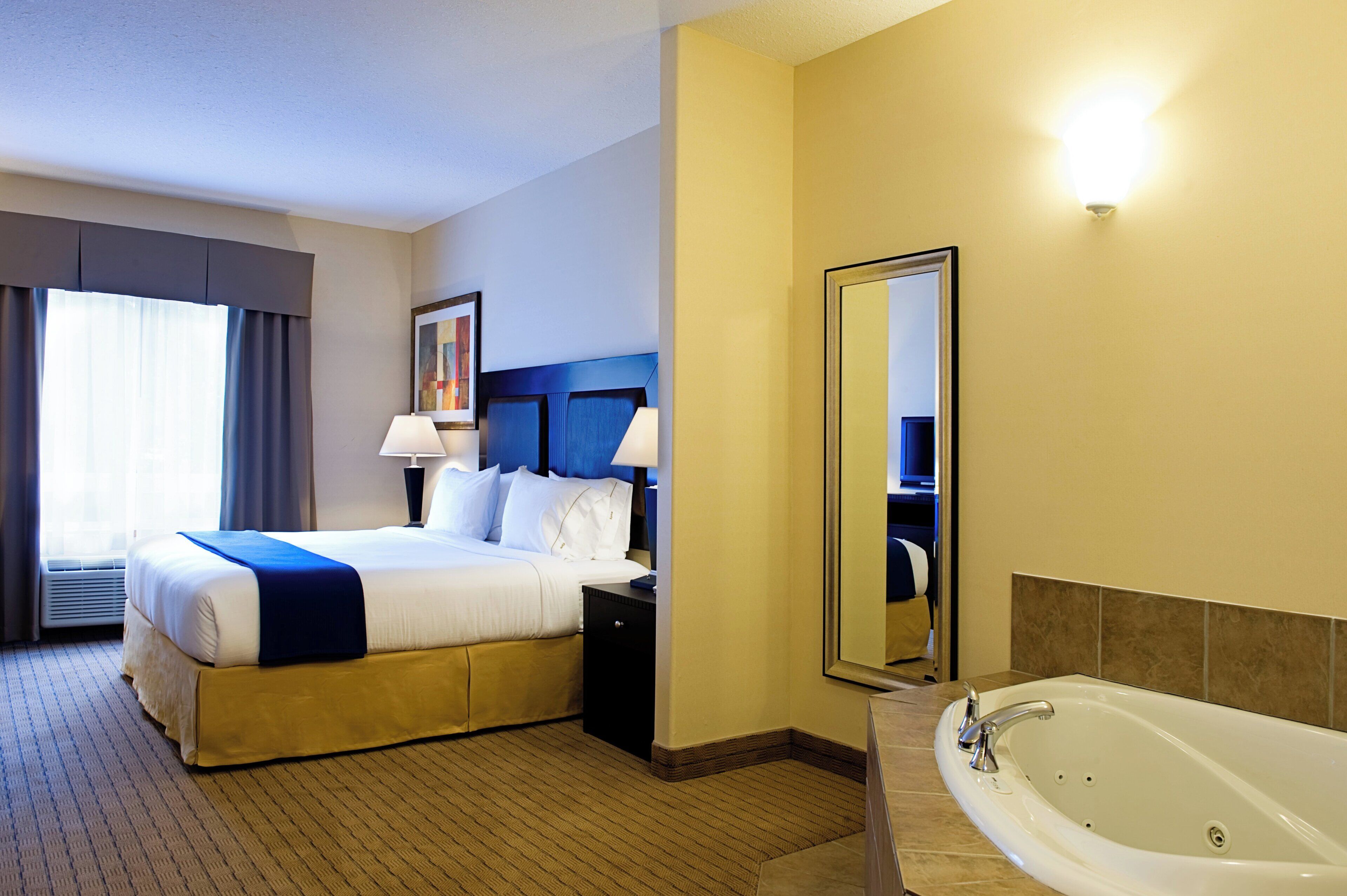 A hotel room with a bed, bedside lamps, a large mirror, a bathroom with a bathtub, and a window with curtains.