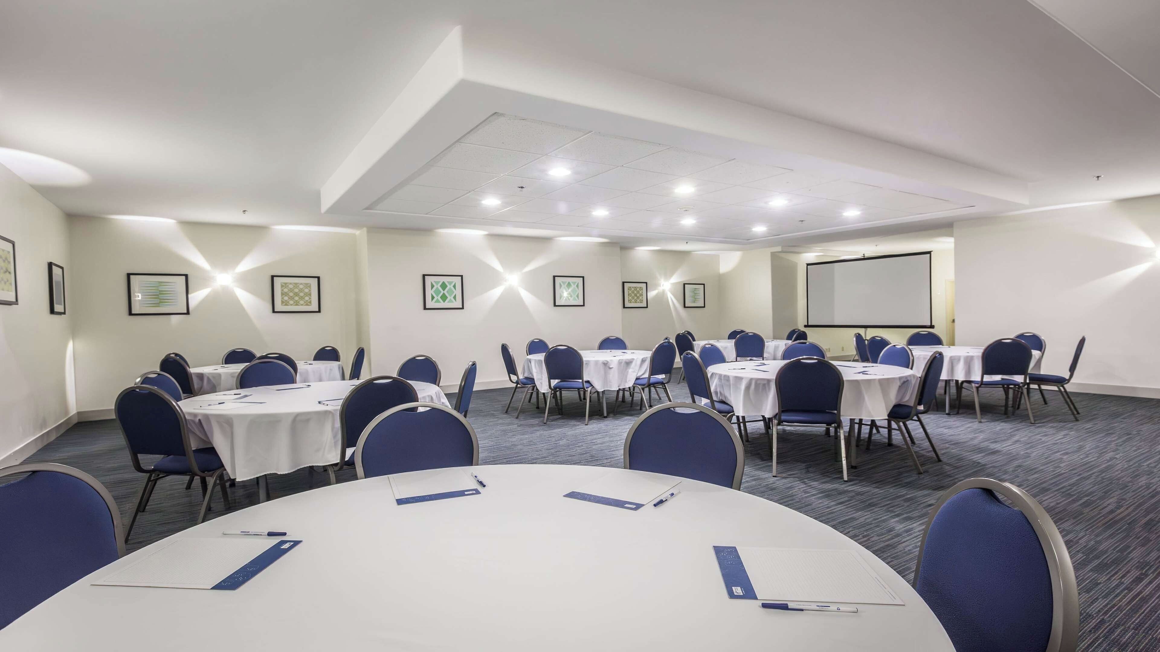 A conference room with multiple round tables, blue chairs, and a projector screen.