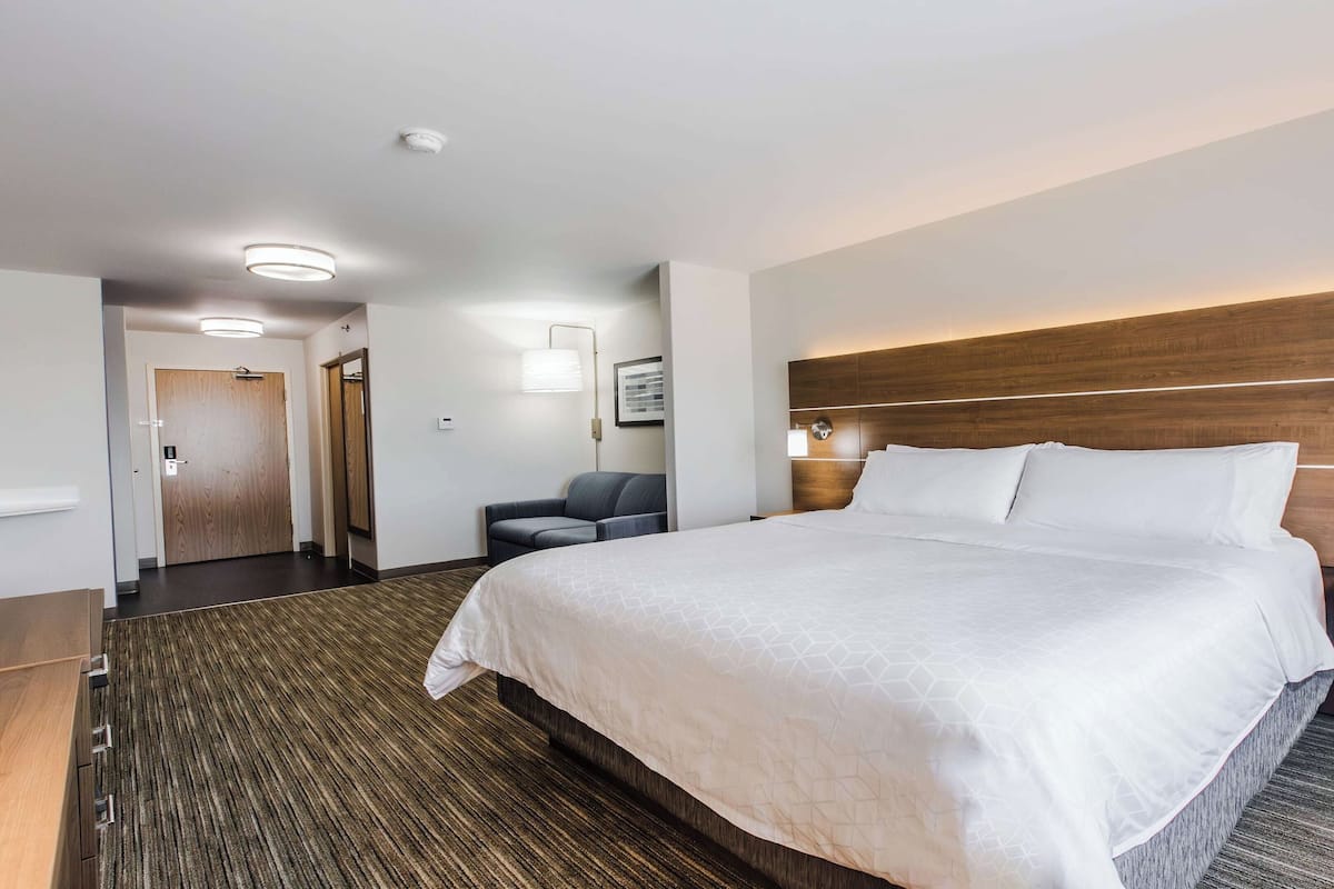 A hotel room with a large bed, a wooden headboard, a nightstand, a seating area with a chair, and a door leading to a hallway.