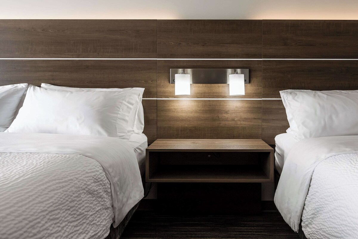 A hotel room with two beds, a wooden headboard, and a nightstand with two wall-mounted lights.