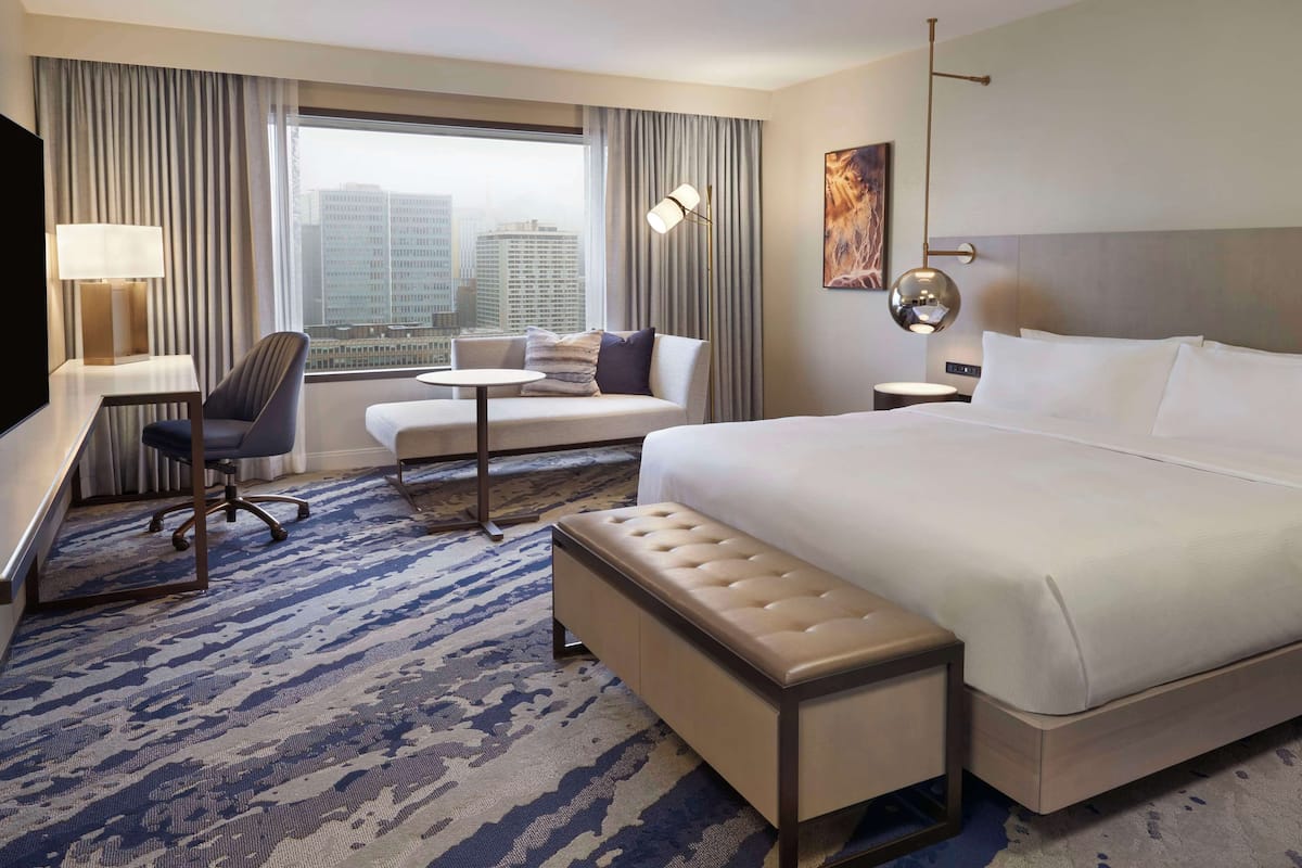 A hotel room with a large bed, a desk with a chair, a small table, and a sofa. There is a city view through the window.