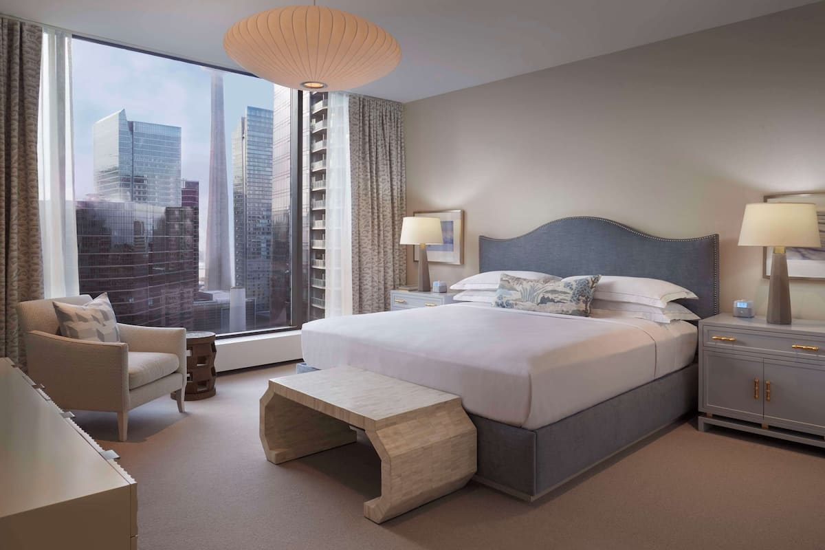 A modern hotel room with a large bed, a seating area with a bench, a nightstand, and a view of the cityscape through large windows.