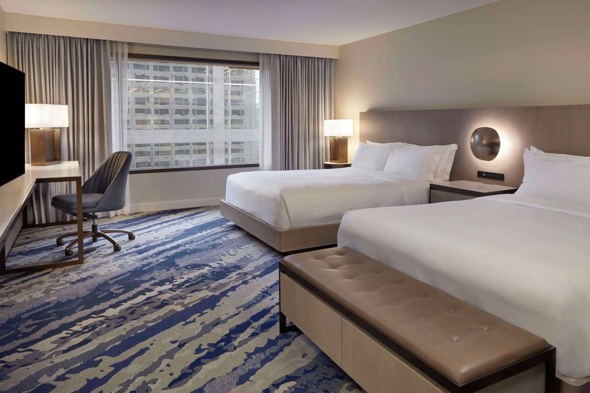 A hotel room with two beds, a desk, a chair, and a large window with curtains.